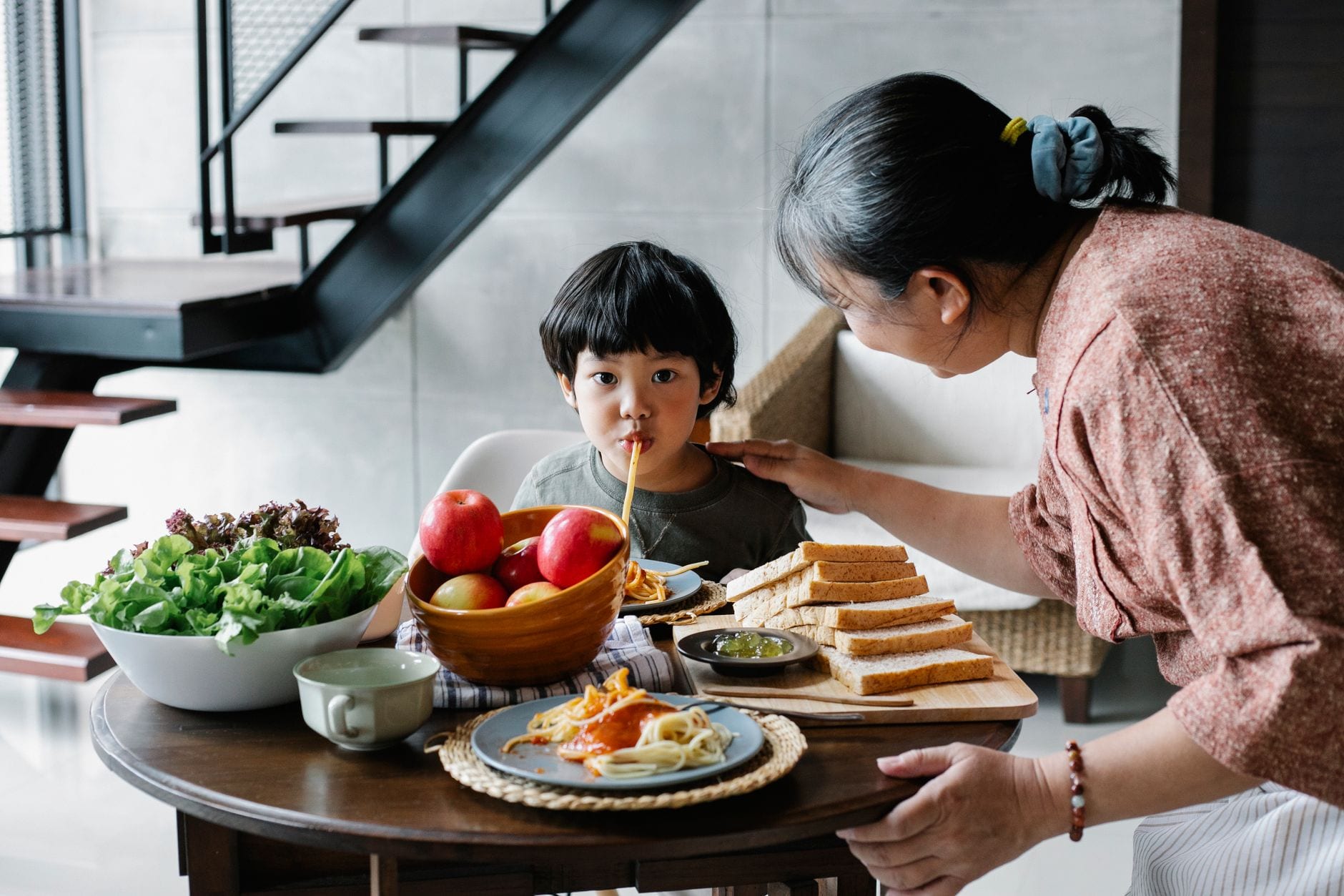 Cute funny Asian boy sucking spaghetti while gathering at dining table with positive grandmother in light kitchen